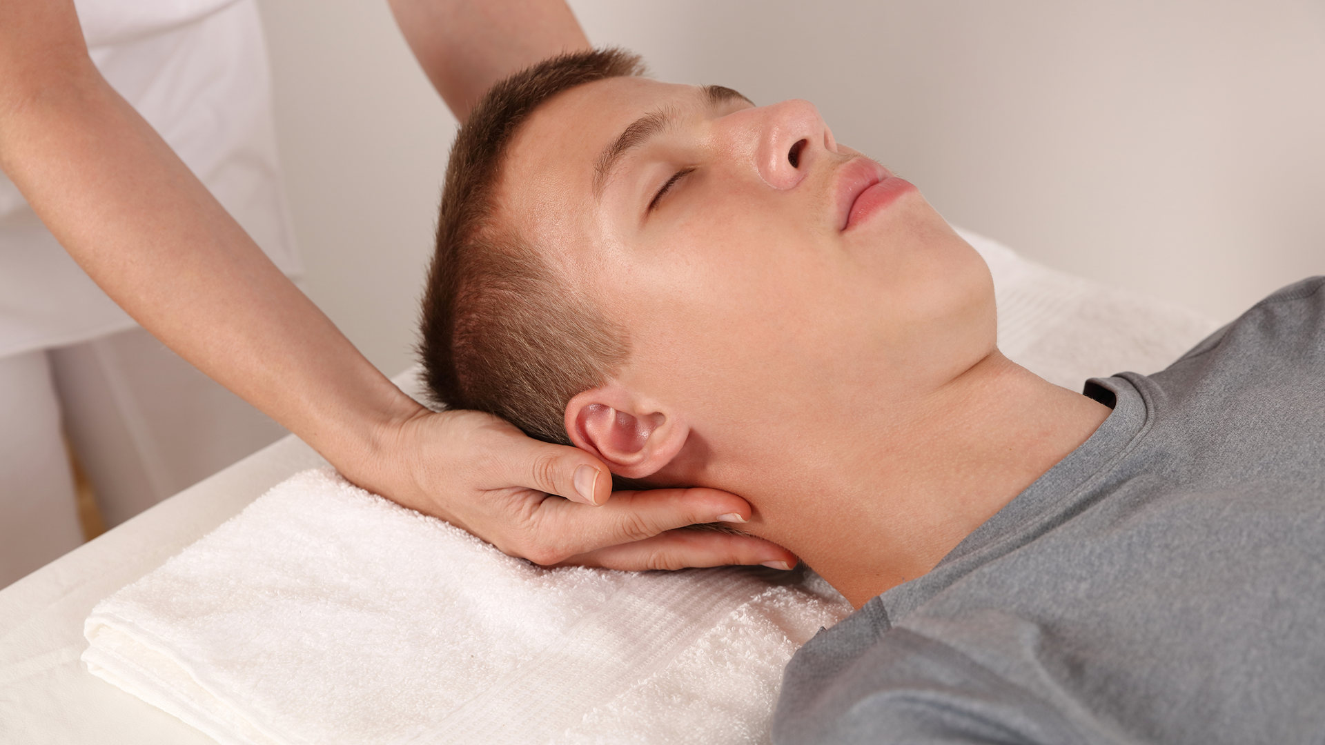 A person lies on a massage table with their eyes closed while receiving a neck massage from a therapist.